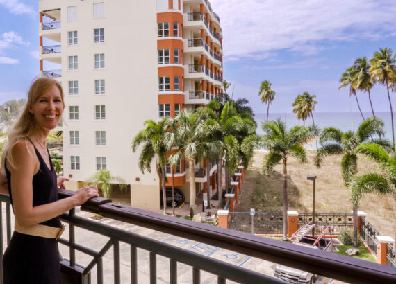 travel blogger michelle hartz smiling and looking at camera on balcony in rincon puerto rico with Caribbean sea and Macor by the sea in background © Joel Hartz