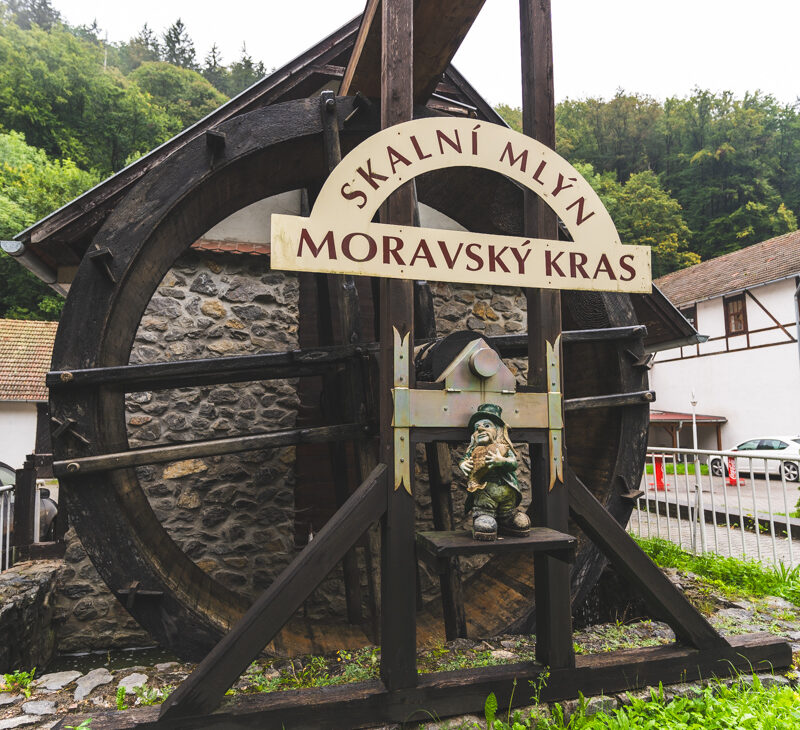 a vertical images of a water wheel with a sign marking the Moravian karst on a cloudy day