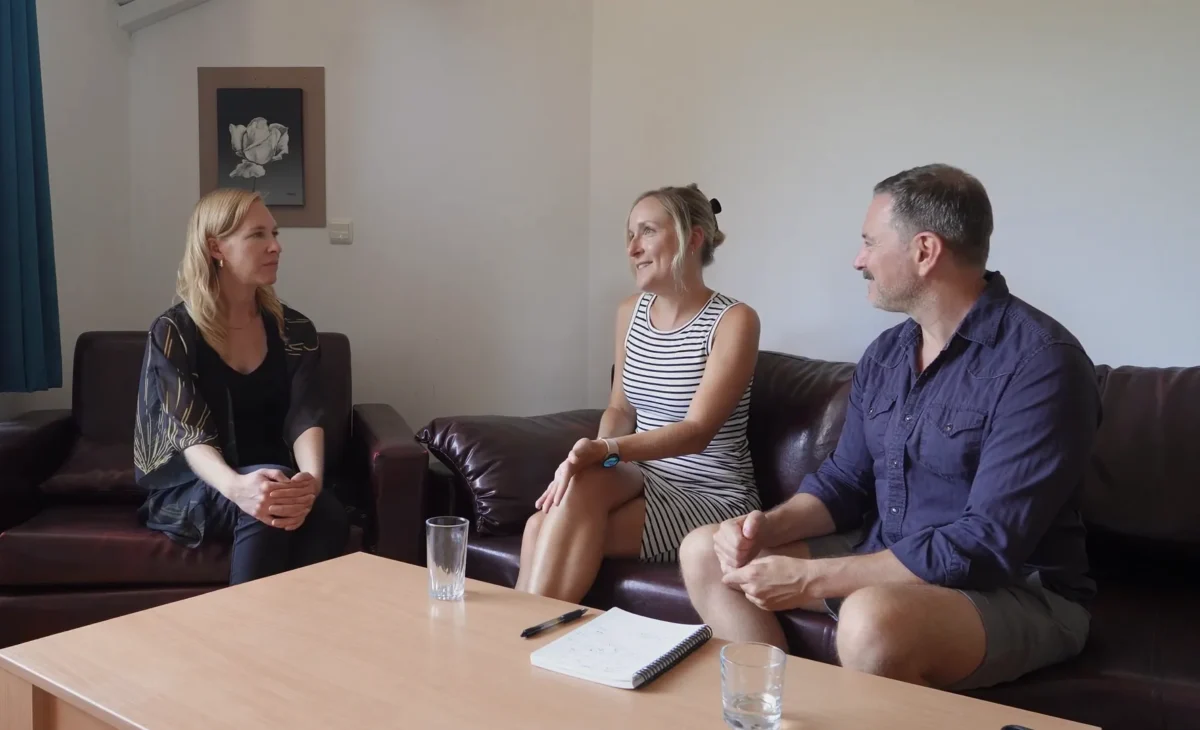 Three people sit on a couch, engaged in a conversation around a coffee table.