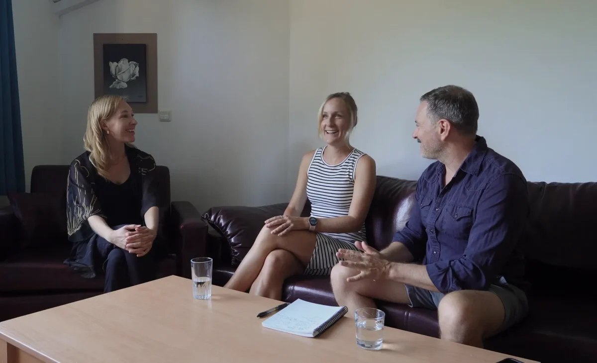 Three people sit on a couch, engaged in a conversation around a coffee table.