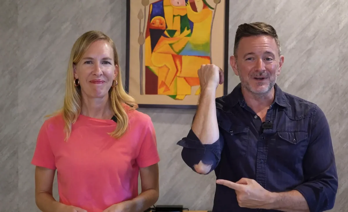 man and woman in front of a picture on the wall. the man is pointing to his torn shirt at the elbow